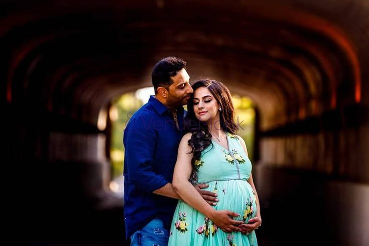 A pregnant woman in a floral aqua dress and a man in a blue shirt embrace in a dimly lit, arched tunnel, with the man gently kissing her cheek as she holds her belly.