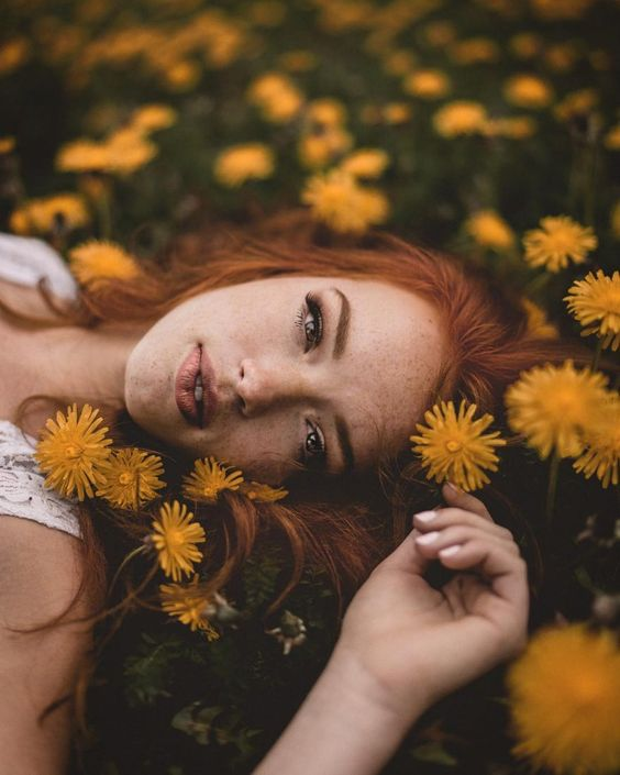 A top-down portrait of a young woman with long, vibrant red hair and freckles lying in a field of yellow dandelions, looking up at the camera with her hand near her face.