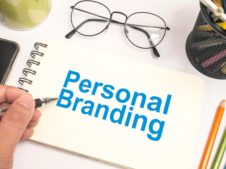 A top-down photograph of a white desk with a spiral notebook, where a hand holds a pen pointing to the words "Personal Branding" written in blue.