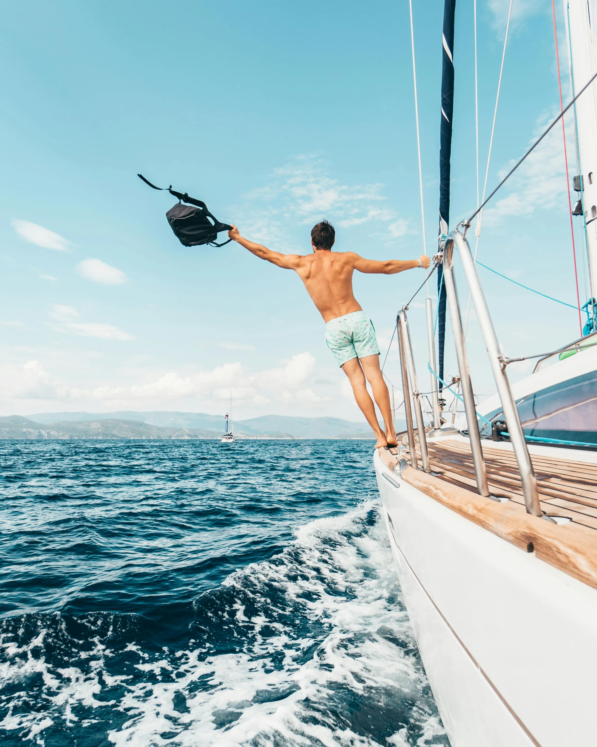 A shirtless man with his back to the camera stands balanced on the bow of a sailboat, leaning out over the blue ocean and holding a black bag, with a distant coastline under a sunny sky.