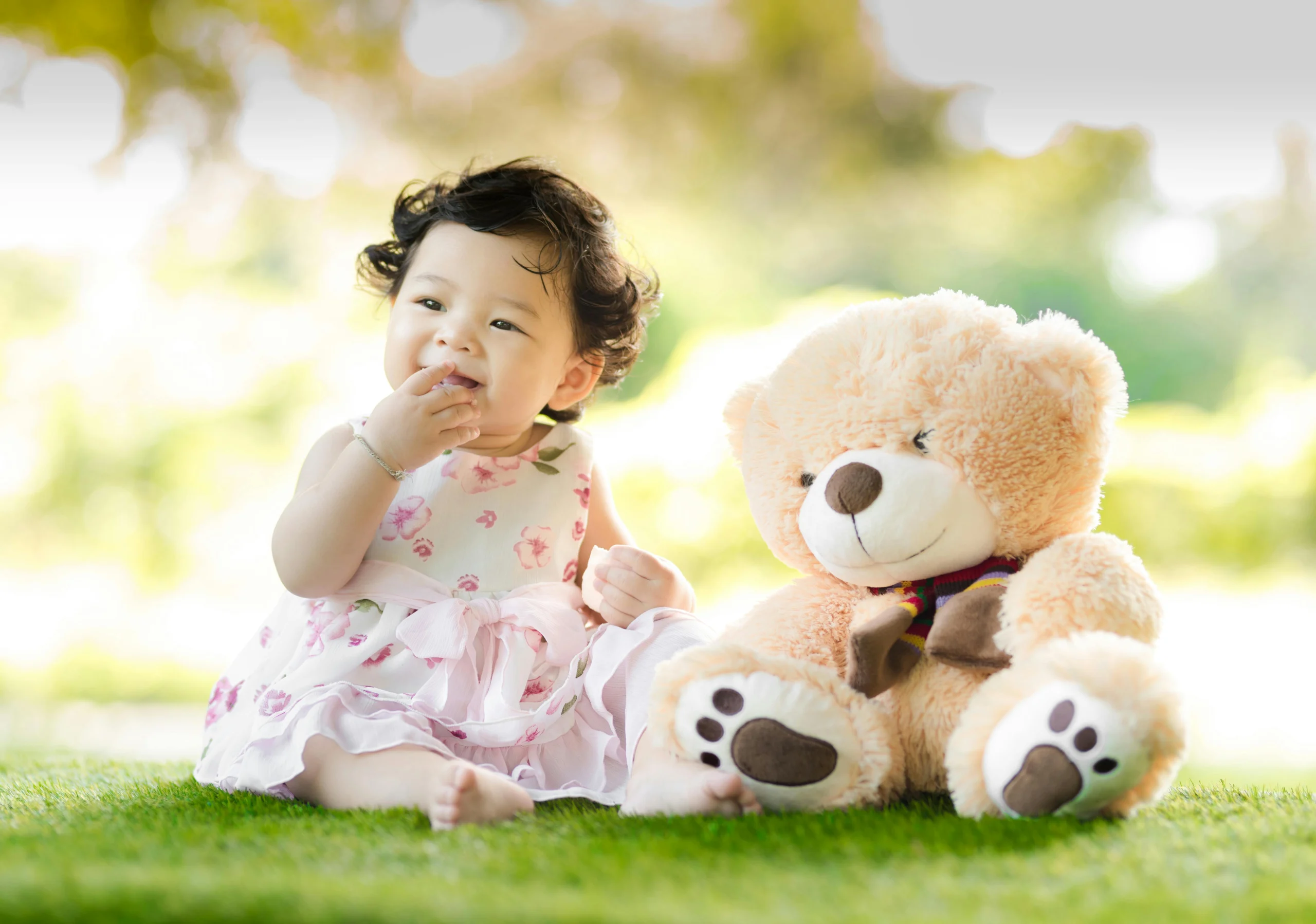 A cute baby girl with curly dark hair, wearing a white and pink floral dress, sits on green grass next to a large teddy bear in a sunlit park.