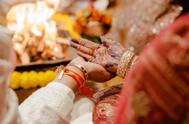 A close-up photograph of a groom holding the henna-adorned hand of his bride during a ritual at an Indian wedding ceremony, with a sacred fire and marigold flowers in the blurred background.