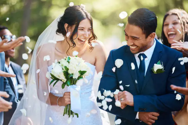 A laughing bride and groom walk through a shower of white flower petals thrown by friends after their wedding ceremony.
