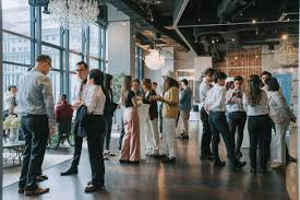 A wide shot of a group of people in business casual attire mingling and talking at a networking event in a modern space with large windows and ornate chandeliers.