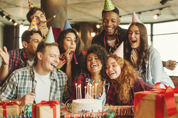A diverse group of seven young adults are happily celebrating a birthday. They are gathered around a table with a lit birthday cake, wearing party hats, blowing noisemakers, and surrounded by gifts and confetti.