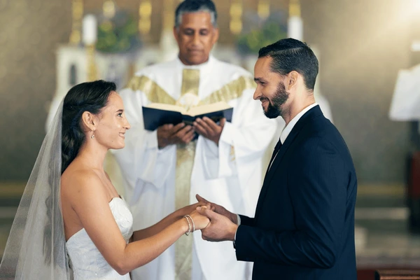 Bride and groom holding hands and smiling at each other during a church wedding ceremony, with an officiant in the background.