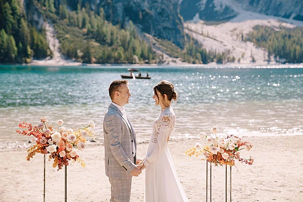 Alt Text: A bride and groom hold hands and smile at each other on a sandy beach during a lakeside wedding ceremony. A turquoise lake with a rowboat and sun glinting on the water, along with large mountains, are in the background. Two floral arrangements with pink and orange roses on metal stands are on either side of the couple.